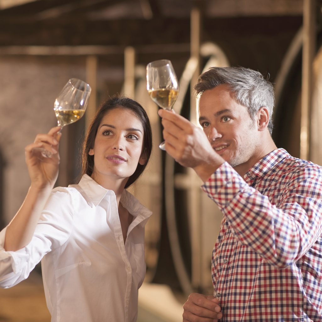 Couple tasting a glass of white wine in a traditional cellar surrounded by wooden barrels.